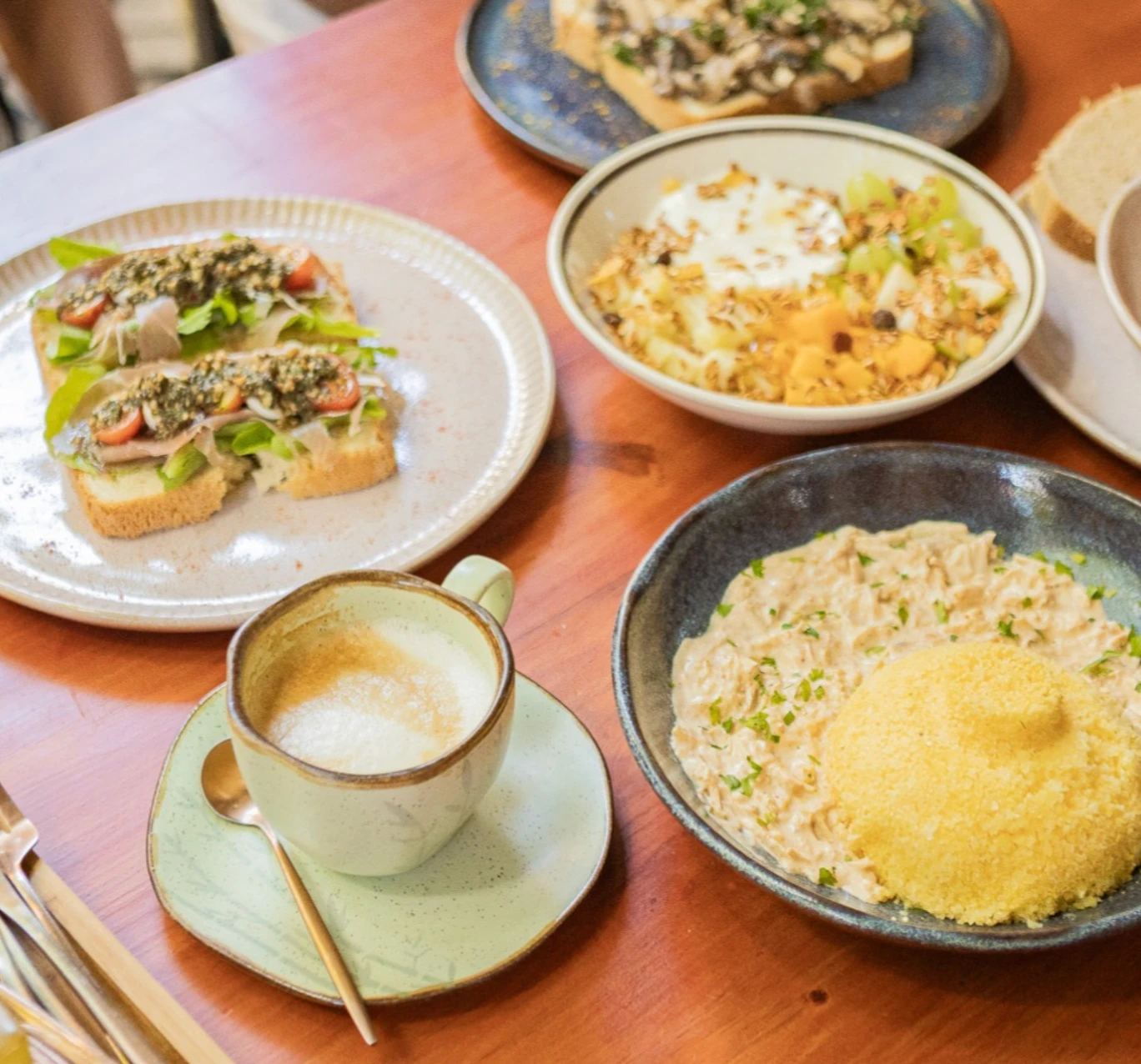 Mesa de café da manhã no Hygge com torrada de parma e pesto, bowl de frutas com granola, cuscuz recheado e cappuccino